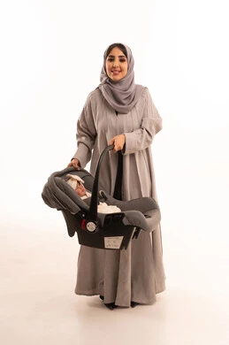 مفهوم الأمومة والرعايه والاهتمام بالأبناء، أهمية الأم ودورها الأساسي في تربية ونشأة الأطفال ، بورترية لامراه عربيه خليجيه سعودية ترتدي العباءة و الحجاب تحمل كرسي طفلتها الرضيعه و تنظر بإيماءات السعادة والحب, الجسم بالطول الكامل، خلفية بيضاء