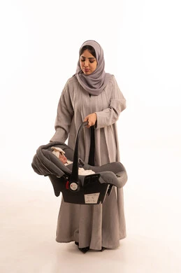 The importance of the mother and her essential role in raising and nurturing children, the concept of motherhood, care, and attention to children, a portrait of an Arab Gulf Saudi woman wearing an abaya and hijab, holding her infant daughter's chair and looking with gestures of happiness and love, full-body, white background.