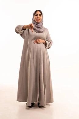 مفهوم الرفض وعدم القبول، بورترية لامراه عربيه خليجيه سعودية حامل ترتدي العباءة و الحجاب تشير بإصبع الابهام للاسفل بإيماءات عدم الاعجاب والتقييم السلبي، الشعور بالانزعاج من شيء ما، صورة الجسم بالطول الكامل, خلفية بيضاء