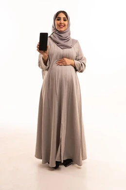 عرض شاشة سوداء فارغة ، بورترية لامراه عربيه خليجيه سعودية حامل ترتدي العباءة و الحجاب تحمل بيدها الهاتف المحمول ، استخدام الأجهزة التقنية الحديثة، الجسم بالطول الكامل، خلفية بيضاء