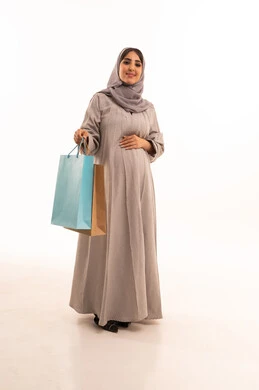 Shopping for supplies and browsing in commercial malls in Saudi Arabia, taking advantage of seasonal offers and discounts, a portrait of a pregnant Arab Gulf Saudi woman wearing an abaya and hijab, holding shopping bags in her hand, looking at the camera with expressions of joy, the positive impact of shopping on women, full-body shot, white background.