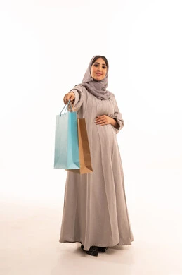 شراء المستلزمات والتبضع من المولات التجارية في السعودية، الاستفادة من العروض الموسمية والتخفيضات،بورترية لامراه عربيه خليجيه سعودية حامل ترتدي العباءة و الحجاب تحمل بيدها اكياس التسوق، النظر الى الكاميرا بتعابير السرور، الأثر الإيجابي للتسوق على السيدات، الجسم بالطول الكامل، خلفية بيضاء