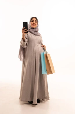 عرض شاشة سوداء فارغه، التسوق عبر الانترنت واقتناء المستلزمات والهدايا , بورترية لامراه عربيه خليجيه سعودية حامل ترتدي العباءة و الحجاب تحمل بيدها هاتف محمول وأكياس التسوق الملونه، الاستفادة من التخفيضات الموسمية, الجسم بالطول الكامل، خلفية بيضاء