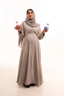 تجهيز مستلزمات الرضيع ، الاهتمام بتغذية ونمو الأبناء، بورترية لامراه حامل عربيه خليجيه سعودية حامل ترتدي العباءة و الحجاب تمسك بيديها رضاعة اطفال زرقاء وورديه ،اختيار اللون المناسب للمولود،  الجسم بالطول الكامل، خلفية بيضاء