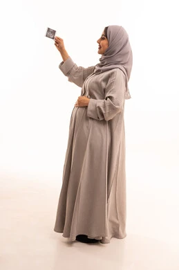 Attention to the health of the fetus, the concept of pregnancy and childbirth, a side portrait of a Saudi Arabian Gulf woman who is pregnant, wearing an abaya and hijab, placing her hand on her belly and holding a sonogram, waiting for the arrival of the newborn, full-body image, white background.