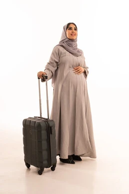 Looking at something, setting off for an enjoyable summer trip, feeling happiness and joy, tourism during the summer vacation, and a portrait of a pregnant Arab Gulf Saudi woman wearing an abaya and hijab holding a travel bag, full-body image, white background.