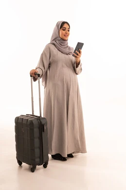 Browsing the internet and social media, tourist flights, chatting with family and friends via mobile, a portrait of a pregnant Arab Gulf Saudi woman wearing an abaya and hijab holding a travel bag and mobile phone in her hands, full-body, white background.
