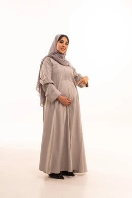تعابير الفرح والسرور، مفهوم الحمل والانجاب، بورتريه لامرأة عربية خليجية سعودية حامل ترتدي العباءة و الحجاب تضع يدها على بطنها و تقوم بالاشارة اليها، انتظار قدوم المولود، النظر إلى الكاميرا بإيماءات الحماس، الاهتمام بصحة الجنين، الجسم بالطول الكامل، خلفية بيضاء