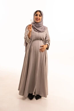 التأكد من نظافة الأسنان بعد تناول الطعام، العناية بالصحة الفموية، الاهتمام بالأناقة والمظهر الخارجي، المحافظة على ابتسامة بيضاء، بورتريه لامرأة عربية خليجية سعودية حامل ترتدي العباءة و الحجاب تمسك فرشاة اسنان، صورة الجسم بالكامل، خلفية بيضاء