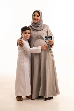 مفهوم العاطفة عند الأمهات، الاطمئنان على مراحل نمو الجنين باستخدام الأشعة فوق الصوتية،  بورترية لامراه حامل عربيه خليجيه سعودية تمسك الاشعه الفوق صوتية برفقة ابنها، الجسم بالطول الكامل، خلفية بيضاء