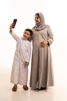 توثيق اللحظات السعيدة عبر تصوير صور السيلفي، مفهوم الحمل و الانجاب، بورترية لولد عربي خليجي سعودي يرتدي الثوب الابيض يمسك الهاتف المحمول برفقة والدته، تعابير السرور و تحقيق الاهداف ، الجسم بالطول الكامل، خلفية بيضاء