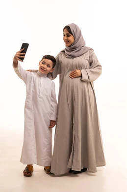 التقاط صور السيلفي التذكارية عبر الجوال، مفهوم الحمل و الانجاب، بورترية لولد عربي خليجي سعودي يرتدي الثوب الابيض يمسك الهاتف المحمول برفقة والدته، تعابير السرور و تحقيق الاهداف ، الجسم بالطول الكامل، خلفية بيضاء