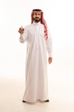 النظر إلى الكاميرا بإيماءات السرور، مركز تجميلي رجالي، مفهوم العناية و الاهتمام بالجمال والمظهر الخارجي، قص و تصفيف الذقن والشارب في صالون الحلاقة، بورتريه لرجل عربي خليجي سعودي  يرتدي الثوب الابيض و الشماغ يحمل بيده المقص، صورة لكامل الجسم، خلفية بيضاء