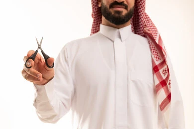 قص و تصفيف الذقن والشارب في صالون الحلاقة، النظر إلى الكاميرا بإيماءات السرور، مركز تجميلي رجالي، مفهوم العناية و الاهتمام بالجمال والمظهر الخارجي، بورتريه  مقرب لرجل عربي خليجي سعودي  يرتدي الثوب الابيض و الشماغ يحمل بيده المقص، خلفية بيضاء