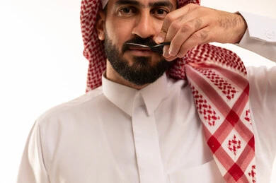 Using scissors in personal care, a men's beauty center, the concept of caring about beauty and appearance, a close-up portrait of a Saudi Arabian Gulf man wearing a white thobe and a ghutrah, trimming and styling his beard and mustache, white background.