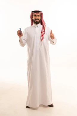 A touch of cleanliness gives freshness and confidence, a mark of excellence and quality, maintaining elegance, skincare, and attention to appearance. A portrait of a Saudi Arabian Gulf man wearing traditional attire and a ghutrah holding a razor and raising his thumb in a like gesture, full-body, white background.