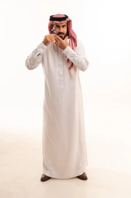 المحافظة على الأناقة والنظافة الشخصية، مفهوم العناية بالبشرة والاهتمام بالمظهر الخارجي، بورتريه لرجل عربي خليجي سعودي يرتدي الثوب التقليدي و الشماغ يقوم بإزالة شعر الوجه باستخدام شفرة الحلاقة الرجالية، الجسم بالطول الكامل، خلفية بيضاء