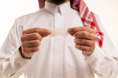 استخدام ماسكات تنظيف المسام ومكونات تجميلية، العناية والاهتمام بنضارة وتحسين البشرة، مفهوم العناية بالنفس، التخلص من الرؤوس السوداء والجلد الميت، بورترية مقرب لرجل عربي خليجي سعودي يرتدي الثوب الابيض و الشماغ يمسك بيدية لصقات الأنف، خلفية بيضاء