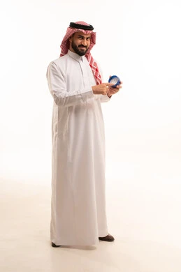 النظر الى الكاميرا بتعابير السرور، استخدام مكونات تجميلية شبابية، مفهوم الإهتمام بنضارة وتحسين البشرة، ترطيب وتنظيف وتفتيح الجلد، روتين العناية بالبشرة، بورتريه لرجل عربي خليجي سعودي يرتدي الثوب الابيض و الشماغ يضع كريم المرطب ، الجسم بالطول الكامل، خلفية بيضاء