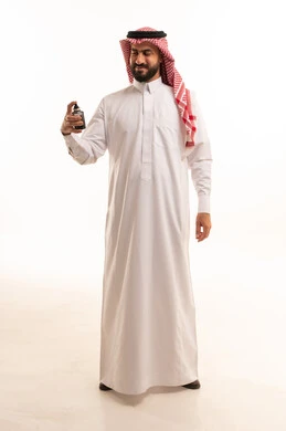 عطورات خليجية سعودية، مفهوم التعطر والتطيب، التسويق لمنتجات العطور, الاهتمام بالمظهر الخارجي, استخدام الزيوت العطرية الطبيعية, بورتريه لرجل عربي خليجي سعودي يرتدي الثوب الابيض و الشماغ يقوم برش العطر, صورة الجسم بالطول الكامل, خلفية بيضاء