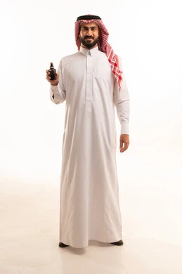 مفهوم التعطر والتطيب، التسويق لمنتجات العطور، عطورات خليجية سعودية, الاهتمام بالمظهر الخارجي, استخدام الزيوت العطرية الطبيعية, بورتريه لرجل عربي خليجي سعودي يرتدي الثوب الابيض و الشماغ يقوم برش العطر, صورة الجسم بالطول الكامل, خلفية بيضاء