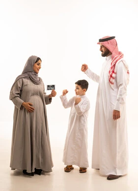 Monitoring the stages of pregnancy and the fetus through the ultrasound device, expressions of achievement and success, the concept of pregnancy and childbirth, a side portrait of a pregnant Arab Gulf Saudi woman wearing an abaya and hijab holding ultrasound images in her hands accompanied by her son and husband, full-body, white background.