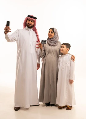 التقاط صور السيلفي عبر الهاتف المحمول، استخدام تقنية السونار لفحص الجنين داخل الرحم، بورترية لامراه حامل عربيه خليجيه سعودية ترتدي العباءة و الحجاب تمسك بيديها اشعه السونار برفقة زوجها و ابنها، مفهوم الحمل و الانجاب، ايماءات السعادة و السرور، الجسم بالطول الكامل، خلفية بيضاء