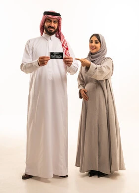 تصوير الجنين بالسونار لمتابعة تطوره، الاشارة باصبع السبابة بايماءات السعادة و السرور، بورترية لرجل عربي خليجي سعودي يرتدي الثوب الابيض و الشماغ يحمل اشعه السونار برفقه زوجته، الجسم بالطول الكامل، خلفية بيضاء