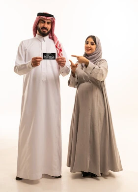 Pointing with the index finger with gestures of happiness and joy, conducting a sonar examination to ensure the health of the child, a portrait of an Arab Gulf Saudi man wearing a white thobe and a shemagh holding a sonar image accompanied by his wife, full-body, white background.