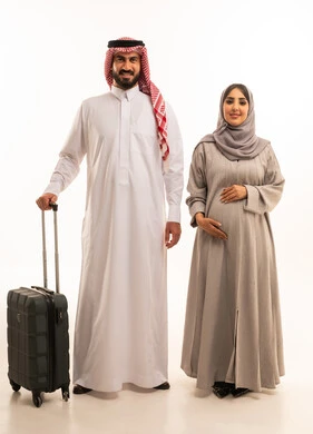 Looking at the camera with expressions of happiness and joy, spending enjoyable times on vacation, enjoying travel and summer trips, a portrait of an Arab Gulf Saudi man wearing a white thobe and a ghutrah holding a travel bag in the company of his wife, the concept of pregnancy and childbirth, a full-body image, white background.