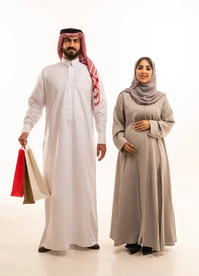 Exploiting seasonal offers and discounts and Black Friday, shopping and taking essential supplies, a portrait of an Arab Gulf Saudi man wearing a white thobe and a shemagh carrying colorful paper bags accompanied by his wife, the concept of pregnancy and childbirth, shopping from commercial malls, gestures of happiness and joy, full-body, white background.