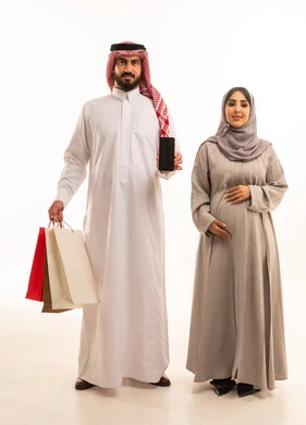 التبضع وأخذ المستلزمات الضرورية،بورترية لرجل عربي خليجي سعودي يرتدي الثوب الابيض و الشماغ يحمل أكياس ورقية ملونه برفقة زوجته, استغلال العروض والتخفيضات الموسمية والجمعة البيضاء، مفهوم الحمل و الانجاب، التسوق من المولات التجارية، ايماءات السعاده و السرور، الجسم بالطول الكامل، خلفية بيضاء
