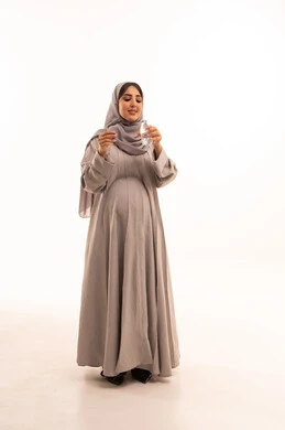 المحافظة على سلامة الجهاز المناعي للجسم، الشعور بالعطش والجفاف، بورتريه لامراة عربيه خليجيه سعودية ترتدي العباءة و الحجاب تقوم بشرب الماء من زجاجة بلاستيكية صغيرة، المداومة على شرب المياه، مفهوم الحمل و الانجاب، الجسم بالطول الكامل، خلفية بيضاء