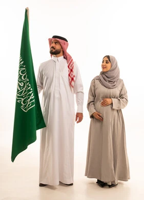 المناسبات والأعياد الوطنية، مشاعر الانتماء للوطن, النظر الى شيء ما، يوم العلم السعودي في 11 مارس, استذكار اليوم الوطني في 23 سبتمبر, بورتريه لرجل عربي خليجي سعودي يرتدي الثوب الابيض و الشماغ يمسك علم المملكة باعتزاز برفقة زوجته, مفهوم الحمل و الانجاب، صورة الجسم بالكامل, خلفية بيضاء