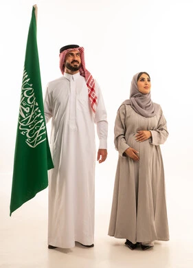 مشاعر الانتماء للوطن, المناسبات والأعياد الوطنية, النظر الى شيء ما، يوم العلم السعودي في 11 مارس, استذكار اليوم الوطني في 23 سبتمبر, بورتريه لرجل عربي خليجي سعودي يرتدي الثوب الابيض و الشماغ يمسك علم المملكة باعتزاز برفقة زوجته, مفهوم الحمل و الانجاب، صورة الجسم بالكامل, خلفية بيضاء