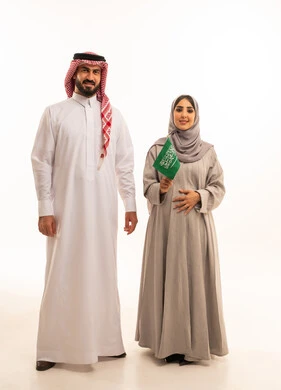 تأسيس الدولة السعودية الأولى 22 فبراير، يوم بدينا 1727 م، الاحتفال بيوم العلم السعودي11 مارس، مفهوم الحمل و الانجاب، بورتريه لإمرأة عربية خليجية سعودية ترتدي العباءة و الحجاب تمسك راية وشعار المملكة بتعابير الفخر برفقة زوجها , الجسم بالطول الكامل، خلفية بيضاء
