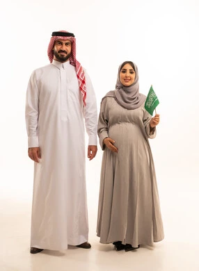 يوم بدينا 1727 م،  ذكرى تأسيس الدولة السعودية الأولى 22 فبراير، الاحتفال بيوم العلم السعودي11 مارس، مفهوم الحمل و الانجاب، بورتريه لإمرأة عربية خليجية سعودية ترتدي العباءة و الحجاب تمسك راية وشعار المملكة بتعابير الفخر برفقة زوجها , الجسم بالطول الكامل، خلفية بيضاء