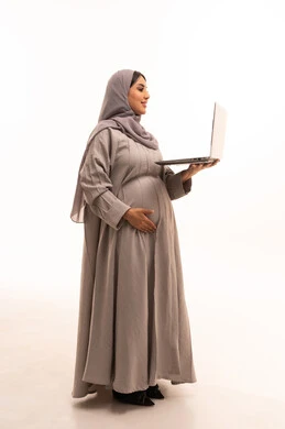استخدام جهاز تقني حديث ومتطور، العمل عن بعد,  مفهوم الحمل و الانجاب، بورتريه من الجانب لامرأة عربية خليجية سعودية محجبة ترتدي العباءة تعمل على جهاز الحاسوب المحمول, انجاز المهام والأعمال, الجسم بالطول الكامل، خلفية بيضاء