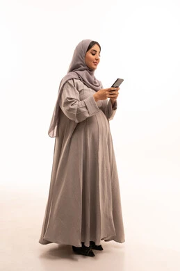 متابعة أخبار وسائل التواصل الاجتماعي باستخدام الجوال، الدردشة والتواصل مع التواصل مع الأصدقاء اونلاين، مفهوم الحمل و الانجاب، بورتريه من الجانب لإمرأة عربية خليجية سعودية ترتدي حجاب وعباءة تمسك بيديها الهاتف المحمول، تعابير وايماءات الفرح والسرور، صورة الجسم بالكامل، خلفية بيضاء