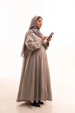 Chatting and communicating with friends online, following social media news using a mobile phone, the concept of pregnancy and childbirth, a side portrait of a Saudi Gulf Arab woman wearing a hijab and abaya holding a mobile phone in her hands, expressions and gestures of joy and happiness, full-body image, white background.