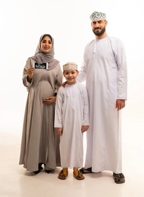 Monitoring the development of the fetus inside the womb through ultrasound, the concept of pregnancy and childbirth, a portrait of an Arab Gulf Omani pregnant woman wearing an abaya and hijab holding a sonogram in her hands accompanied by her son and husband, full-body, white background.