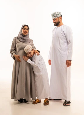 ايماءات السعادة و السرور،  مفهوم الترابط الأسري ، الشوق والارتباط بالجنين القادم، بورترية لولد عربي خليجي عماني يرتدي الدشداشة و الكمه يضع رأسه على بطن والدته برفقة ابيه ، الجسم بالطول الكامل، خلفية بيضاء