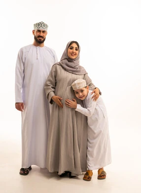 الشوق والارتباط بالجنين القادم، بورترية لولد عربي خليجي عماني يرتدي الدشداشة و الكمه يضع رأسه على بطن والدته برفقة ابيه ، مفهوم الترابط الأسري ، ايماءات السعادة و السرور، الجسم بالطول الكامل، خلفية بيضاء