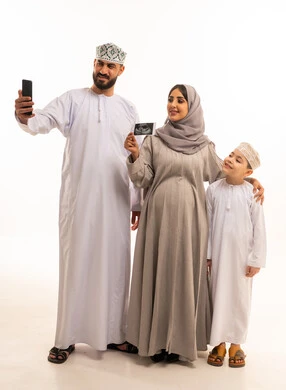 التقاط صور السيلفي عبر الهاتف المحمول، استخدام تقنية السونار لفحص الجنين داخل الرحم، بورترية لامراه حامل عربيه خليجيه عمانية ترتدي العباءة و الحجاب تمسك بيديها اشعه السونار برفقة زوجها و ابنها، مفهوم الحمل و الانجاب، ايماءات السعادة و السرور، الجسم بالطول الكامل، خلفية بيضاء