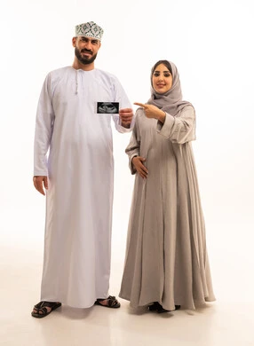 Ultrasound imaging of the fetus to monitor its development, pointing with the index finger with gestures of happiness and joy, a portrait of an Arab Gulf Omani man wearing a dishdasha and a kumma holding the ultrasound image alongside his wife, full-body, white background.