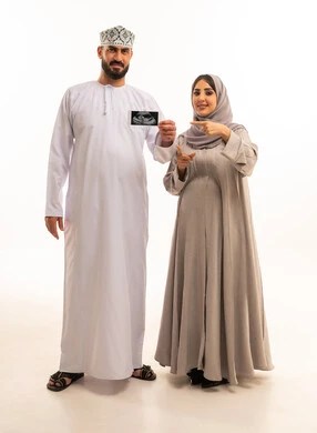 الاشارة باصبع السبابة بايماءات السعادة و السرور، إجراء فحص السونار للاطمئنان على صحة الطفل، بورترية لرجل عربي خليجي عماني يرتدي الدشداشة و الكمه يحمل اشعه السونار برفقه زوجته، الجسم بالطول الكامل، خلفية بيضاء