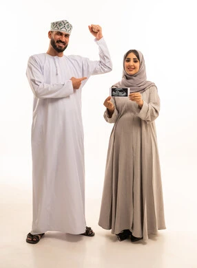 إجراء فحص السونار للاطمئنان على صحة الطفل، بورترية لامراه حامل عربيه خليجيه عمانية ترتدي العباءة و الحجاب تمسك بيديها اشعه السونار برفقه زوجها ، الاشارة بايماءات السعادة و السرور، الجسم بالطول الكامل، خلفية بيضاء