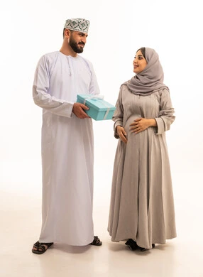 مشاركة وتقديم الهدايا في الأعياد والمناسبات العائلية, بورتريه لرجل عربي خليجي عماني يرتدي الدشداشة و الكمة يقدم لزوجته صندوق الهديه، مفهوم الحمل و الانجاب، الألفة والمحبة بين الزوجين, الجسم بالطول الكامل، خلفية بيضاء