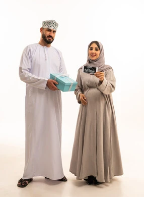 تقديم الهدايا في العيد والمناسبة الخاصة، بورتريه لرجل عربي خليجي عماني يرتدي الدشداشة والكمة يحمل صندوق الهدية برفقه زوجته ، امراه حامل عربيه خليجيه عمانية تحمل بيديها اشعه السونار ، مفهوم الحمل و الانجاب، الجسم بالطول الكامل، خلفية بيضاء