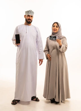 استخدام السونار للاطمئنان على الطفل, بورترية لرجل عربي خليجي عماني يرتدي الدشداشة و الكمة يقوم بعرض شاشة سوداء عبر الهاتف المحمول ,  امراه  حامل خليجيه عمانية ترتدي العباءة و الحجاب تمسك بيديها الاشعه الصوتية، مفهوم الحمل و الانجاب, الجسم بالطول الكامل, خلفية بيضاء